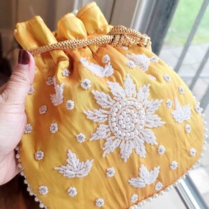 May include: A yellow drawstring bag with white pearl embellishments and floral designs. The bag has a gold drawstring closure.
