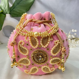 May include: A pink velvet drawstring bag with gold and pearl embellishments. The bag has a paisley design and a large, round, gold-toned embellishment in the center.