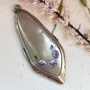 May include: A silver locket pendant with a floral design. The locket is oval shaped and has a detailed border. The flowers are painted in shades of purple and green.
