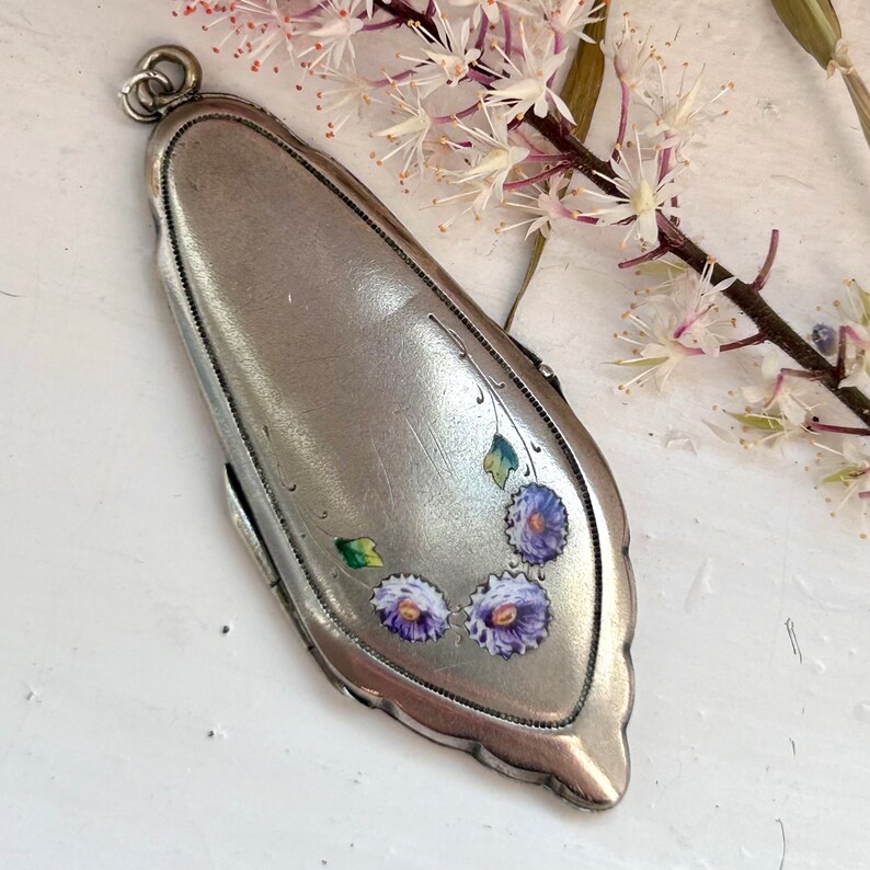 May include: A silver locket pendant with a floral design. The locket is oval shaped and has a detailed border. The flowers are painted in shades of purple and green.