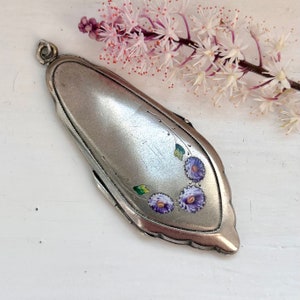 May include: A silver locket with a floral design. The locket has a hinged lid and is decorated with three purple flowers and green leaves.