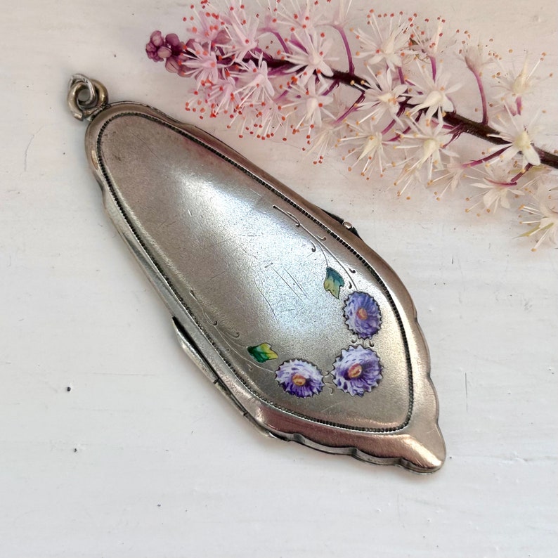 May include: A silver locket with a floral design. The locket has a hinged lid and is decorated with three purple flowers and green leaves.