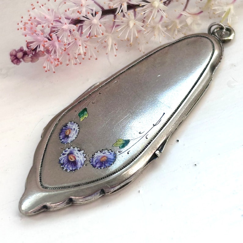 May include: A silver locket with a floral design. The locket is oval shaped and has a hinged opening. The flowers are painted in shades of purple and green.