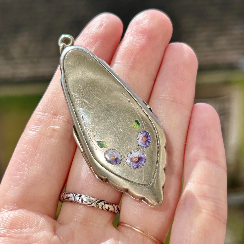May include: Silver locket pendant with a floral design. The locket is oval shaped and has a silver finish. The floral design is painted in purple, green, and white enamel.
