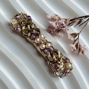 Puede incluir: Una pinza para el pelo dorada con un diseño delicado y ornamentado. La pinza está adornada con cuentas moradas y blancas.