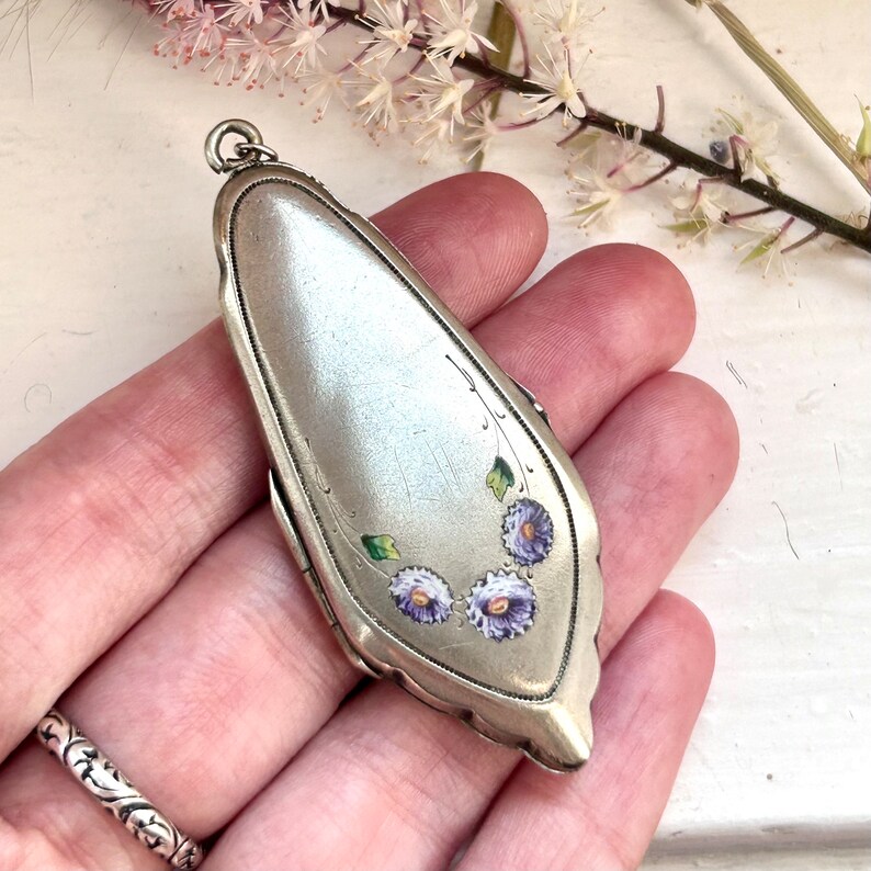 May include: A silver locket pendant with a floral design. The locket is oval shaped and has a hinged lid. The flowers are painted in shades of purple and green.