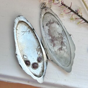 May include: A silver locket with a hinged opening, featuring three small, dark purple stones set in the interior. The locket is slightly tarnished and has a worn, antique appearance.