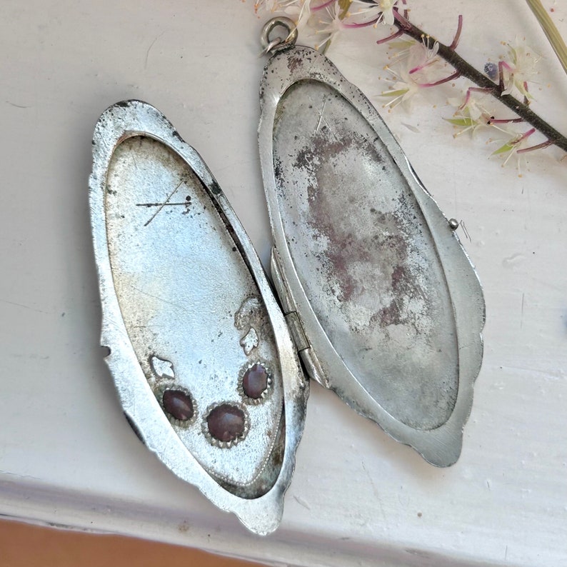 May include: A silver locket with a hinged opening, featuring three small, dark purple stones set in the interior. The locket is slightly tarnished and has a worn, antique appearance.