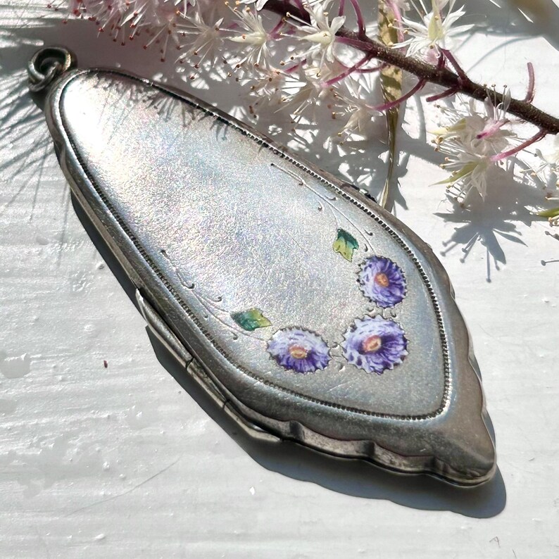 May include: A silver locket with a floral design. The locket is oval shaped and has a hinged lid. The flowers are purple and green and are painted on the front of the locket.