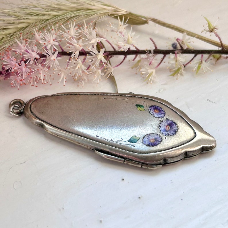 May include: A silver compact with a floral design. The flowers are purple and green and are painted on the front of the compact.