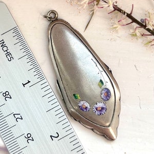 May include: A silver locket pendant with a floral design. The locket is decorated with hand-painted purple and green flowers. The locket is approximately 2 inches long.