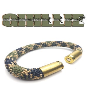 Puede incluir: Un brazalete de camuflaje hecho de paracord y una vaina de bala gastada. La vaina de bala es de latón y tiene un acabado dorado.