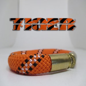 May include: Orange, black and white paracord bracelet with a brass bullet casing. The bracelet is woven in a chequered pattern.
