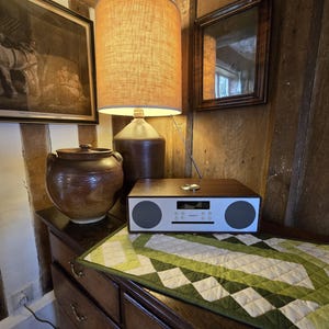 Może przedstawiać: Drewniane radio z białym wyświetlaczem i dwoma głośnikami stoi na stole z zielonym i białym pikowanym bieżnikiem. Na stole stoi r&oacute;wnież brązowy ceramiczny dzbanek i lampa z brązową podstawą i beżowym kloszem.