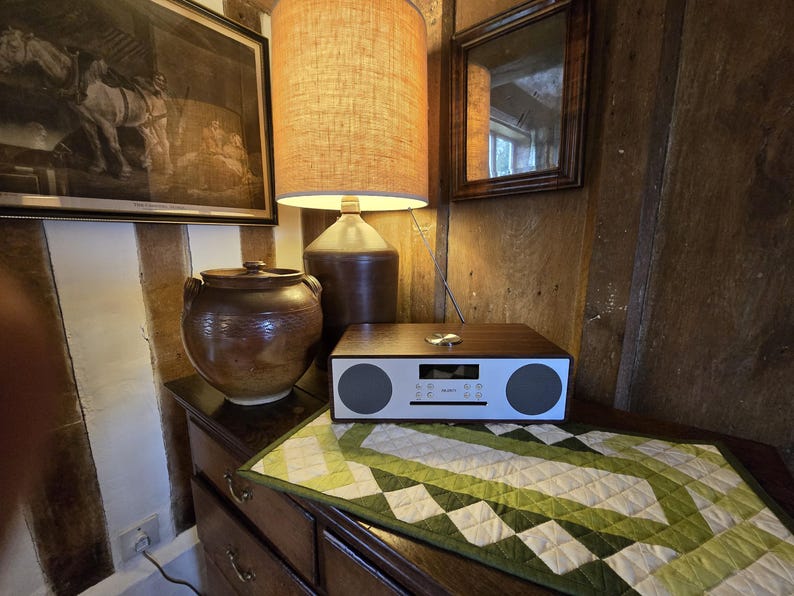 Może przedstawiać: Drewniane radio z białym wyświetlaczem i dwoma głośnikami stoi na stole z zielonym i białym pikowanym bieżnikiem. Na stole stoi r&oacute;wnież brązowy ceramiczny dzbanek i lampa z brązową podstawą i beżowym kloszem.