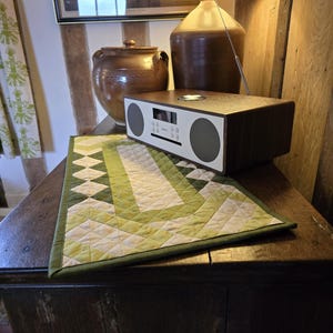 Może przedstawiać: Drewniany st&oacute;ł z zielonym i białym pikowanym bieżnikiem. Na stole stoi brązowy ceramiczny dzbanek i brązowe radio ze srebrną tarczą. W tle znajduje się lampa z brązowym abażurem.