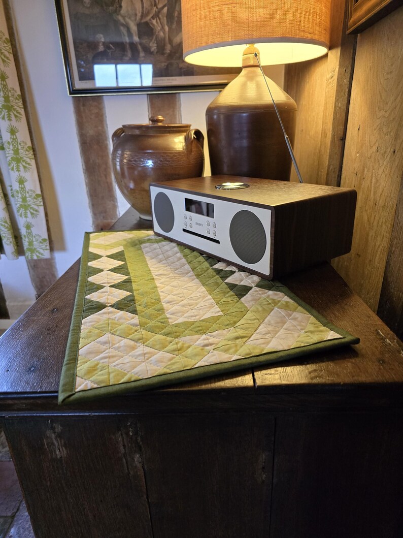 Może przedstawiać: Drewniany st&oacute;ł z zielonym i białym pikowanym bieżnikiem. Na stole stoi brązowy ceramiczny dzbanek i brązowe radio ze srebrną tarczą. W tle znajduje się lampa z brązowym abażurem.