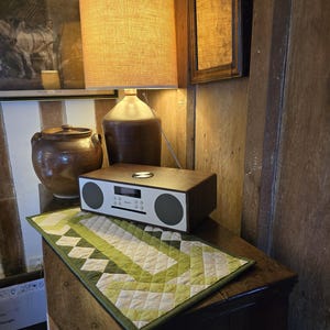 Może przedstawiać: Drewniane radio z białym wyświetlaczem i zielono-białym pikowanym bieżnikiem na drewnianym stole. Radio ma wyświetlacz cyfrowy i pokrętło do regulacji głośności. Bieżnik ma geometryczny wz&oacute;r.