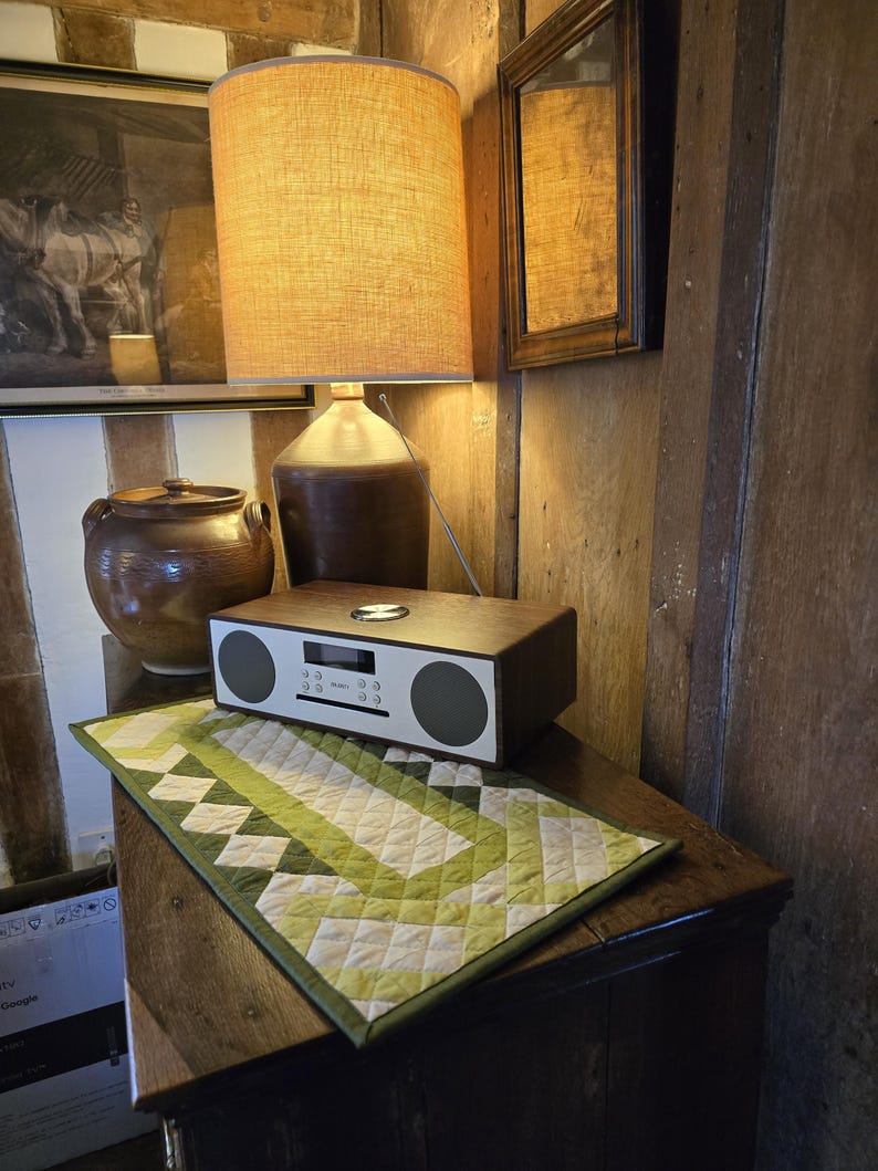 Może przedstawiać: Drewniane radio z białym wyświetlaczem i zielono-białym pikowanym bieżnikiem na drewnianym stole. Radio ma wyświetlacz cyfrowy i pokrętło do regulacji głośności. Bieżnik ma geometryczny wz&oacute;r.