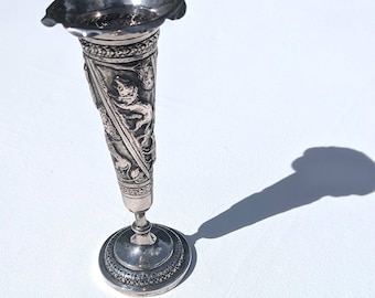 May include: An antique silver bud vase with a flared top and detailed relief carvings. The vase has a round base and a slender, conical body with ornate designs. The vase is approximately 15 cm tall.