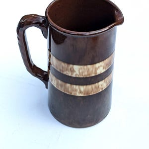 May include: A brown ceramic pitcher with a handle and a spout. The pitcher has two horizontal bands of cream and brown around the middle. The pitcher is on a white surface.