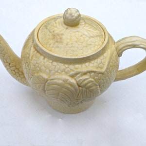 May include: A yellow ceramic teapot with a floral design. The teapot has a lid with a knob on top. The teapot is on a white background.