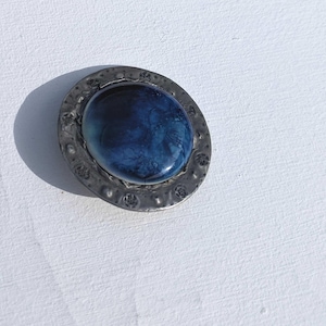 May include: A silver brooch with a blue and black glass cabochon set in a textured metal frame.