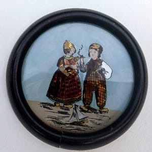 Puede incluir: Una obra de arte vintage, enmarcada y redonda, que representa a dos figuras con trajes tradicionales, una sosteniendo un pequeño animal y la otra fumando en pipa. Un pequeño velero está representado en el agua. El marco es negro.