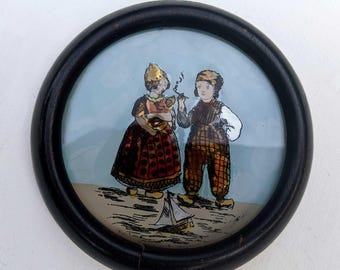 Pintura vintage de temática holandesa sobre vidrio, con iridiscencia, en un pequeño marco redondo de madera negra (requiere presilla para colgar), circa 1920-1930.