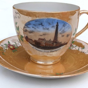 Puede incluir: Un juego de taza y platillo vintage con acabado dorado. La taza presenta una escena pintada de una torre y edificios con el texto "A Present From Blackpool". Diseños florales y de volutas adornan la taza y el platillo.