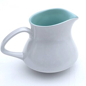 May include: A white ceramic jug with a rounded body and a curved handle. The interior of the jug is a light teal colour, contrasting with the exterior. The jug has a spout for easy pouring.