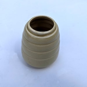 May include: A small, cream-coloured ceramic vase with a ribbed design. The vase is round and has a wide opening at the top.