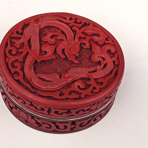 May include: A round, red lacquered box with intricate carved dragon and floral designs. The lid features a detailed dragon motif, while the sides showcase floral patterns. A silver band accents the box's midsection, adding a touch of elegance.