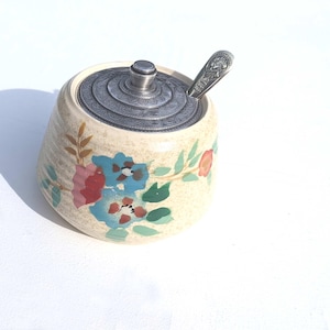 Könnte beinhalten: Eine weiße Zuckerdose aus Keramik mit einem Blumendesign in Blautönen, Rosa und Grün. Die Dose hat einen silbernen Deckel mit einem dekorativen Muster und wird mit einem passenden silbernen Löffel geliefert.