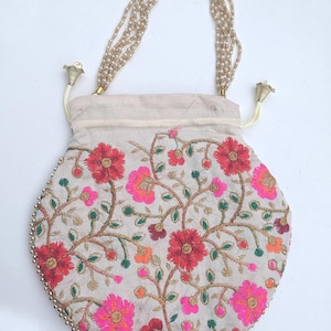 May include: A white round drawstring bag with a floral embroidery design. The bag has a beaded handle and gold-colored metal accents.