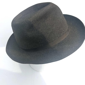Może przedstawiać: Ciemnoszary kapelusz typu fedora ze strukturalną główką i szerokim, lekko zakrzywionym rondem. Kapelusz posiada pasującą ciemnoszarą wstążkę rypsową otaczającą podstawę główki, co zapewnia schludny i jednolity wygląd całego nakrycia głowy.