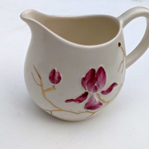 Puede incluir: Una jarra de crema de cerámica blanca con un diseño floral dorado y rosa. La jarra tiene un cuerpo redondeado y un asa.