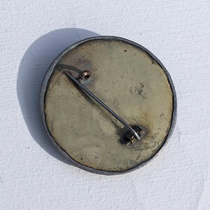 May include: The back of a round, silver brooch with a brass pin and clasp. The brooch has a textured, aged surface.