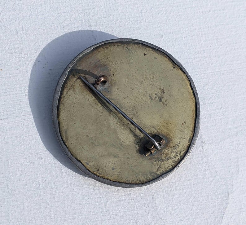 May include: The back of a round, silver brooch with a brass pin and clasp. The brooch has a textured, aged surface.
