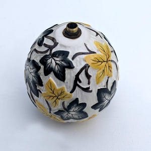 Può includere: Un tappo per bottiglia in ceramica bianca con un design di foglie nere e gialle. Il tappo per bottiglia ha una parte superiore in ottone.