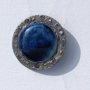 May include: A round, blue glass cabochon set in a silver-toned metal frame with a textured border. The cabochon has a swirling, marbled pattern.