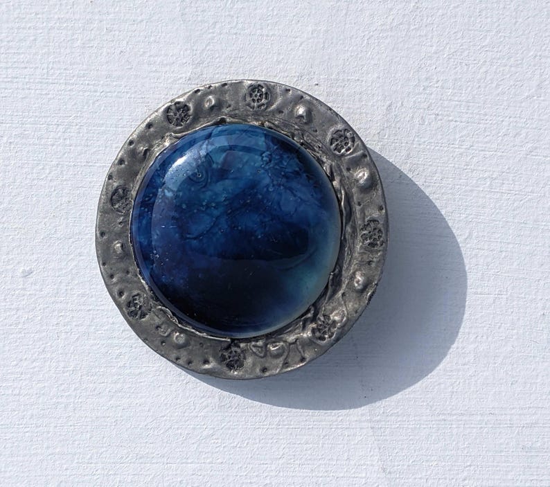 May include: A round, blue glass cabochon set in a silver-toned metal frame with a textured border. The cabochon has a swirling, marbled pattern.