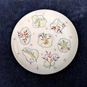 Puede incluir: Un plato de cerámica blanco con un diseño floral azul y amarillo. El plato presenta hojas y bayas estilizadas en tonos rojos, verdes y amarillos.