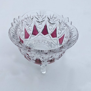 Könnte beinhalten: Eine klare, geschliffene Glasschale mit gewelltem Rand und drei kleinen Füßen. Die Schale hat ein geometrisches Muster mit rubinroten Akzenten. Die Schale wird wahrscheinlich zum Servieren oder Ausstellen verwendet.