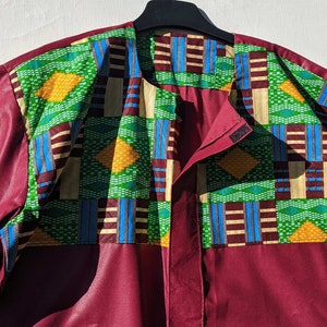 May include: A burgundy shirt with short sleeves and a colourful patterned front panel. The panel features green, yellow, blue, and brown geometric designs. The shirt is hanging on a black hanger.