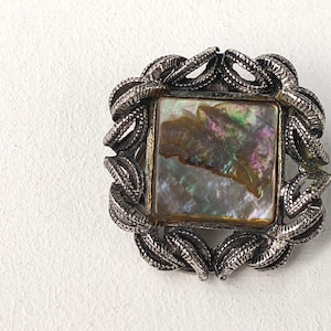 May include: A square brooch with a silver-toned, textured frame. The center features a square iridescent abalone shell. The frame has a decorative, rope-like design. The brooch is set against a white background.