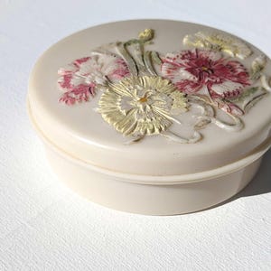 Könnte beinhalten: Eine weiße Keramik-Schmuckdose mit einem erhabenen Blumendesign. Die Blumen sind rosa, weiß und grün.