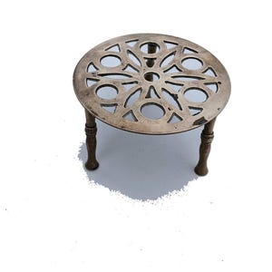May include: A silver-coloured, round trivet with a decorative openwork design. The trivet has three short, turned legs. The openwork pattern features a symmetrical arrangement of circles and petal-like shapes. It is designed to protect surfaces from hot cookware.