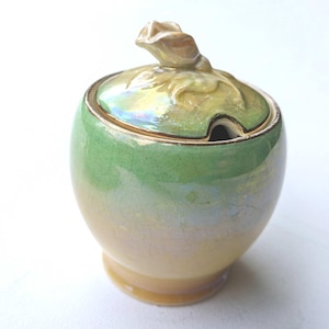 Puede incluir: Un azucarero de cerámica vintage con tapa y una pequeña abertura para una cuchara. El cuenco es un degradado de verde y amarillo con un borde dorado. La tapa está rematada con una flor decorativa.