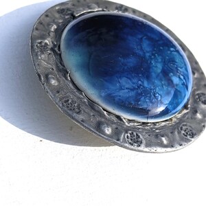 May include: A round pewter brooch with a blue and white glass cabochon in the center. The pewter has a textured surface and is decorated with raised designs.